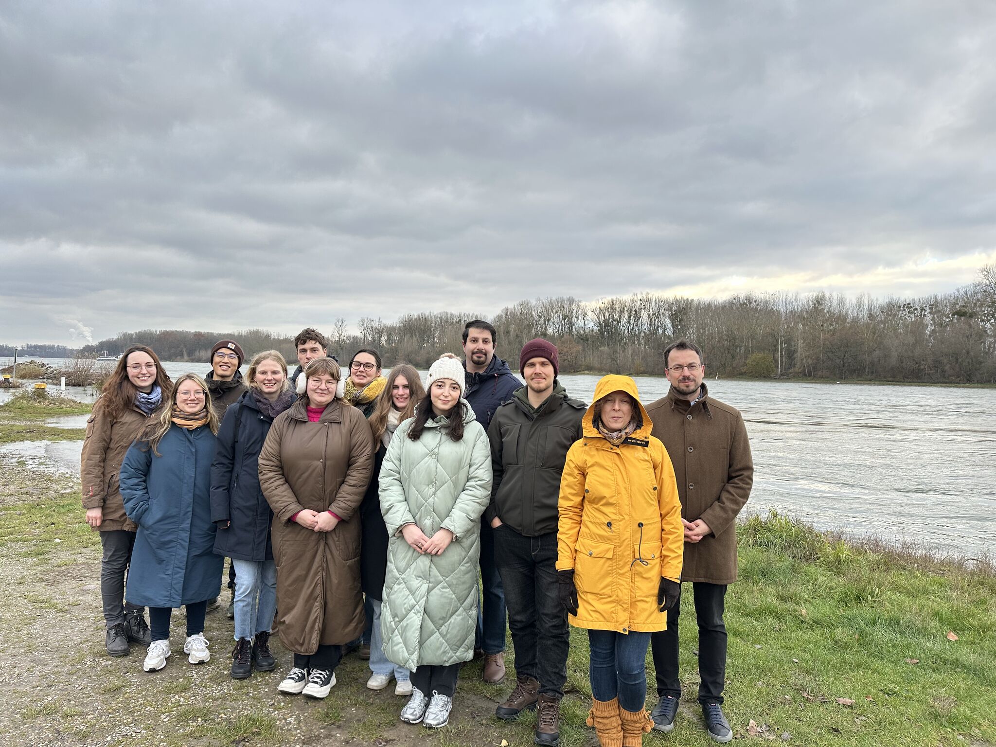 Gruppe von Personen steht am Ufer eines Flusses bei bedecktem Himmel, alle in Winterkleidung.
